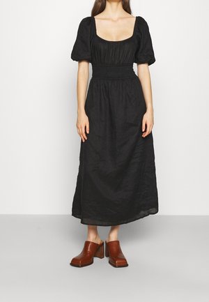 Kobieta ubrana w czarną sukienkę midi z bufkami na rękawach i marszczonym pasem oraz brązowe buty na obcasie bez zapięć, stojąca na szarej podłodze.