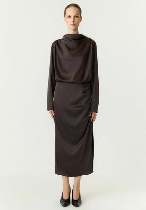Femme debout portant une robe longue en satin marron foncé à manches longues avec un col montant et des escarpins noirs à bout pointu sur un fond clair uni.