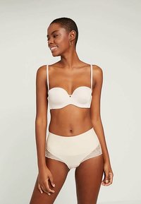 Leende kvinna med kort hår som bär en beige bandeau-bh och högmidjade underkläder med spetsdetaljer mot en neutral bakgrund.