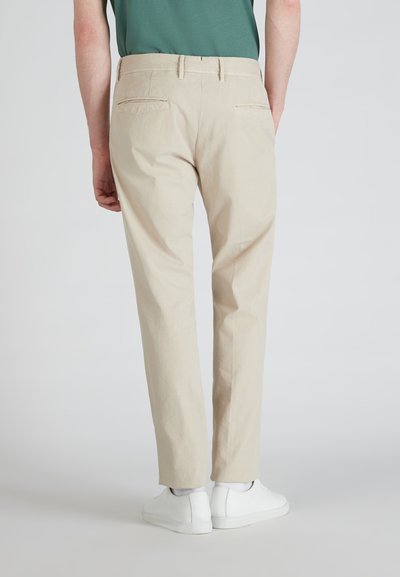 Beige chinos met een slim fit, voorzien van twee achterzakken en subtiele stikdetails, gecombineerd met witte sneakers op een lichte achtergrond.