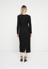 Vestido preto de mangas longas em tecido texturizado com riscas horizontais. Comprimento médio com uma fenda na parte de trás, combinado com sandálias pretas de tiras.