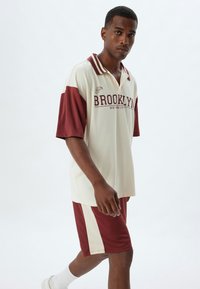 Biała koszulka polo z krótkim rękawem z burgundowymi akcentami i napisem "BROOKLYN"; w połączeniu z pasującymi burgundowymi szortami. Gładki, lekki materiał.
