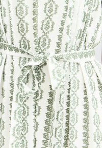 Tissu blanc avec des rayures florales verticales vertes noué en nœud à la taille, créant des plis froncés autour de la ceinture.