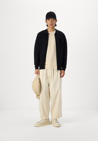 Crna jakna sa zatvaračem, bež majica, široke hlače u boji krem, bež torba. Model nosi crni šešir i krem tenisice. Minimalistički dizajn.