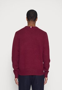 Pull en tricot bordeaux avec texture côtelée, manches longues et encolure en V. Accent rayé notable au niveau du col arrière.