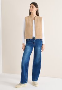 Femme portant un gilet couleur sable à fermeture éclair sur une chemise blanche à manches longues, un jean large bleu et des baskets beige, debout sur un sol blanc.