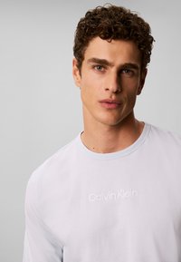 Jeune homme aux cheveux bruns bouclés portant une chemise blanche Calvin Klein sur un fond gris clair uni.