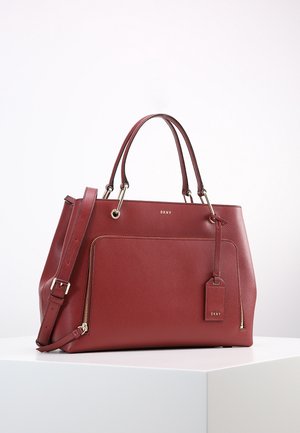 Bolso DKNY de cuero rojo con asas dobles, correa de hombro ajustable, bolsillo delantero con cremallera y etiqueta de marca adjunta sobre superficie blanca.