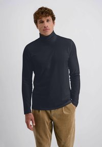 Schwarzes Rollkragen-Shirt mit langen Ärmeln, glatter Textur und tailliertem Design, kombiniert mit hellbraunen Cordhosen. Minimale Markenkennzeichnung sichtbar.