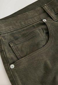 Gros plan d'un pantalon en jean vert foncé montrant la poche avant, la poche à monnaie, la passants de ceinture et les rivets métalliques.