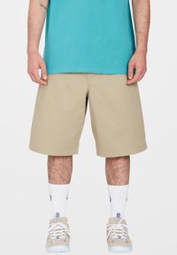 Beige shorts gemaakt van lichtgewicht stof, met een losse pasvorm en omgeslagen zoom. Gecombineerd met een teal t-shirt en witte sportkousen.