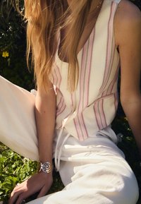 Lightweight, striped sleeveless top in cream with pink and purple lines. White pants. Decorative spiral bracelet on wrist. Green grass background.