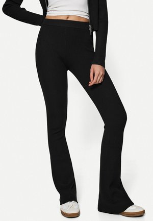 Vrouw draagt zwarte ribgebreide wijde broek met hoge taille, wit cropped top, zwart jack met rits en witte sneakers met bruine zolen.