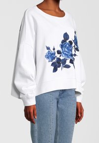 Sudadera blanca de gran tamaño con rosas bordadas en azul, combinada con jeans de un azul claro, modelo mostrado desde el cuello hasta la mitad del muslo, fondo neutro.
