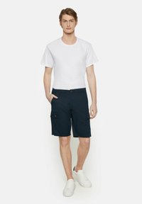 Shorts cargo bleu marine avec deux poches latérales, fermeture à bouton et un t-shirt blanc ajusté. Portés avec des baskets blanches, tissu lisse, design décontracté.