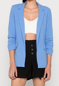 Vaaleansininen blazer, jossa on muotoiltu istuvuus, liehuva kaulus ja kolme neljännesosa hihaa, yhdistetty valkoiseen ribbattuun toppiin ja mustiin räätälöityihin shortseihin.