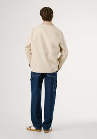 Hombre con chaqueta beige, jeans oscuros con bolsillos laterales y zapatillas blancas, de pie de espaldas contra un fondo neutro.