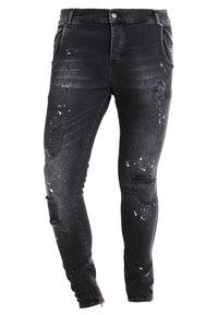Zwarte denim jeans met meerdere versleten plekken, witte verfspatdetails, slim fit en ritsaccenten bij de enkels.