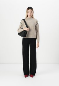 Donna che indossa un maglione beige, pantaloni neri a gamba larga, scarpe rosse e porta una borsa a tracolla nera, in piedi su uno sfondo bianco.