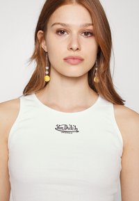 Baltas įribbintas marškinėliai be rankovių, kuriame yra juodas "Von Dutch Originals" logotipas krūtinėje. Modelis turi ilgus plaukus ir dėvi ryškiai geltonas šypsenėlių auskarus.