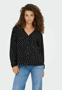 Zwarte blouse met een patroon, witte bloemenontwerpen, V-hals, lange mouwen en knoopsluiting. Gepaarde met lichtblauwe jeans.