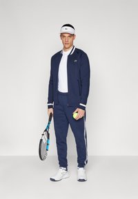 Fato de treino atlético azul-marinho com detalhes brancos, combinado com uma viseira branca. O modelo segura uma raquete de ténis e uma bola de ténis amarela.