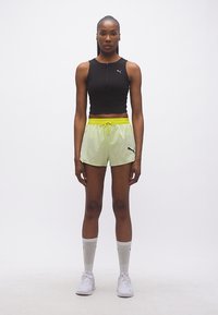 Γυναίκα που στέκεται κοιτάζοντας μπροστά, φορώντας μαύρο αμάνικο crop top, ανοιχτοπράσινα σορτς με κίτρινη μέση, άσπρες κάλτσες και άσπρα αθλητικά παπούτσια.