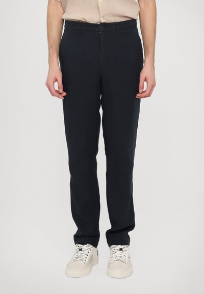 Marineblauwe linnen broek met een rechte pasvorm, voorzien van een ritssluiting en knoopsluiting, gecombineerd met beige sneakers en een lichtgekleurde shirt.