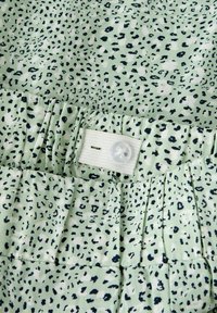 Tissu vert clair à imprimé léopard noir, doté d'une taille élastique réglable avec un bouton blanc et une boutonnière.