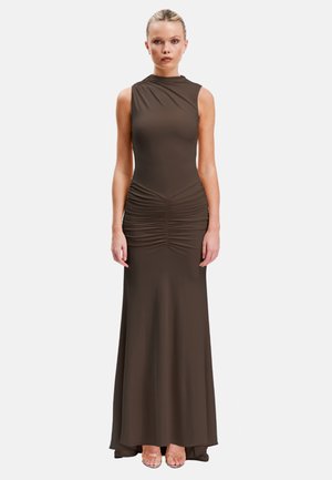 Femme portant une robe maxi sans manches marron foncé froncée avec un col montant et des talons transparents à bouts ouverts, debout face à un fond blanc.