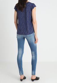 Donna che indossa una blusa floreale navy a maniche corte, jeans skinny blu chiaro e scarpe slip-on nere, in piedi con la schiena rivolta verso una sfondo bianco puro.