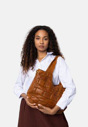 Donna con capelli ricci, in camicia bianca e gonna marrone, che tiene una grande borsa di pelle marrone davanti a uno sfondo bianco.