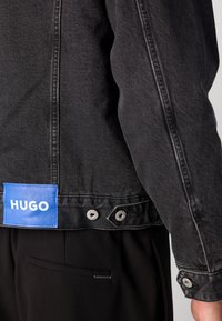 Chaqueta de mezclilla negra que presenta un parche azul con el logo de HUGO en la parte posterior, botones de presión plateados y costuras blancas contrastantes a lo largo de las costuras.