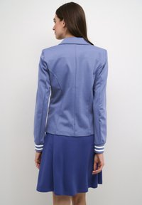 Giacca blu con un design aderente, dotata di colletto e polsini a strisce. Indossata sopra un vestito blu coordinato con un orlo a campana.