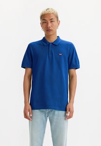 Polo shirt azul em uma mistura de algodão leve, com colarinho clássico, mangas curtas e um pequeno logótipo vermelho no peito.