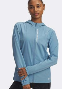 Mujer con una chaqueta con cremallera y capucha de color azul claro de Under Armour, de pie con los brazos cruzados contra un fondo blanco liso.