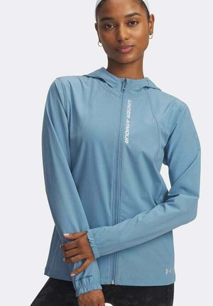 Woman wearing a light blue Under Armour zip-up jacket with a hood, standing with arms crossed against a plain white background.