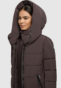 Brązowa ocieplana kurtka z dużym, regulowanym kapturem, kieszeniami na zamek i zatrzaskami. Tekstura jest gładka z pikowanymi sekcjami.