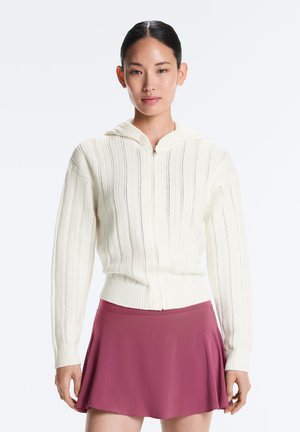 Jonge vrouw met donker haar in een staart, die een witte gebreide hoodie met ritssluiting en een korte bordeauxrode rok draagt tegen een effen achtergrond.