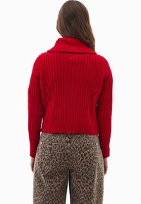 Czerwony sweter prążkowany z szerokim kołnierzem, zestawiony z brązowymi dżinsami w panterkę. Widoczne detale teksturowanej tkaniny i skróconego fasonu.