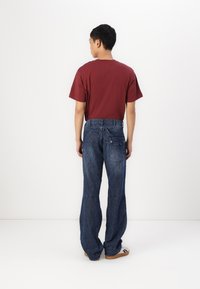 Maroon T-shirt, blue denim pants with a loose fit and distinctive stitching, paired with brown sandals; viewed from the back.