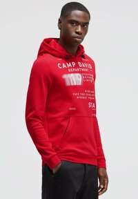 Red hoodie featuring a drawstring, front pocket, and bold white text with branding. Smooth texture, fitted shape, long sleeves, and ribbed cuffs.