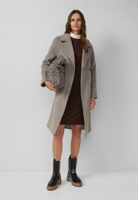 Manteau en laine gris clair avec un col pointu, associé à une robe marron à mi-mollet. Bottines noires à semelles épaisses. Tenant un sac à pois.