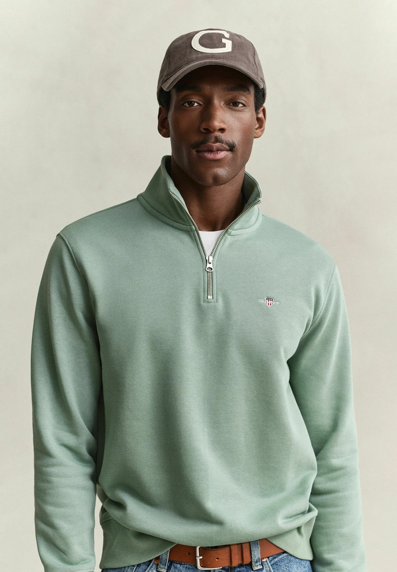 Man with mustache wearing light green quarter-zip pullover, brown belt, and brown cap with letter G on plain background.