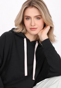 Hanorac negru cu șnururi roz, croială relaxată, mansete ribbate și textură moale. Modelul are părul deschis la culoare și își sprijină mâna pe bărbie.