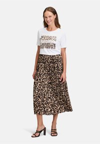 Witte t-shirt met een luipaardprint, gecombineerd met een zwarte en bruine plisseer maxi rok. Zwarte haksandalen accentueren de outfit.