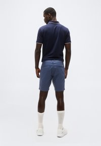 Άνδρας που στέκεται με την πλάτη, φοράει μπλε πουκάμισο πόλο, μπλε σορτς, λευκές ψηλές κάλτσες και λευκά αθλητικά σε απλό φωτεινό φόντο.