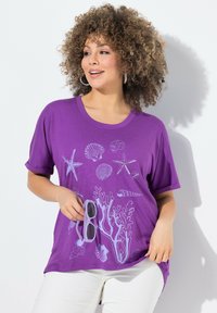 Camiseta de algodón morada con mangas cortas, que presenta impresiones gráficas blancas de temática oceánica con conchas y corales en el frontal. El modelo sostiene unas gafas de sol.
