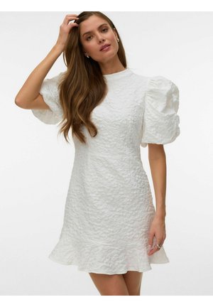 Femme portant une robe blanche texturée avec manches courtes bouffantes et ourlet à volants, touchant ses cheveux d'une main, sur fond uni.