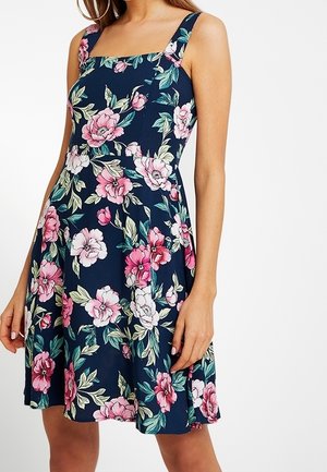 Femme portant une robe bleu marine sans manches avec larges bretelles et motif floral rose et blanc, debout devant un fond blanc uni.